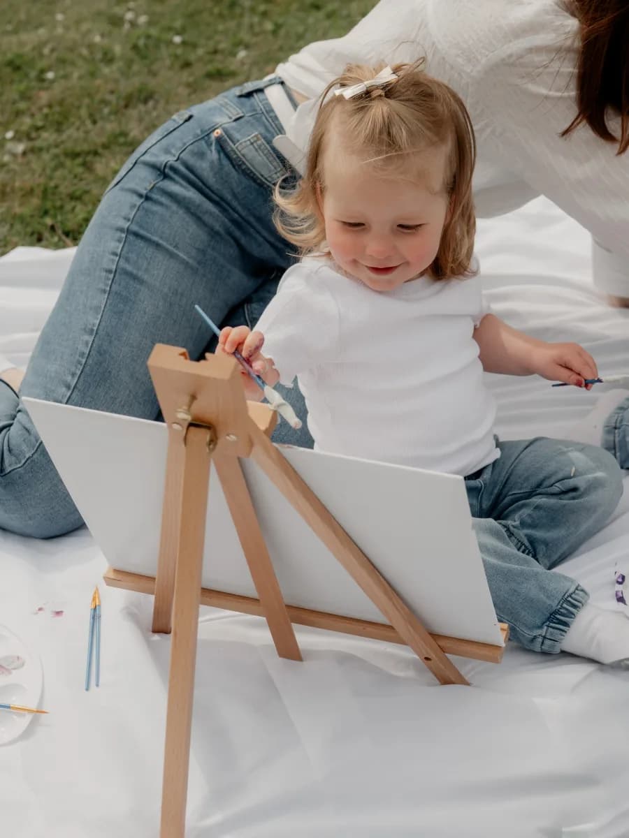 A young child in a white t-shirt sitting on a white sheet with an adult, holding a wooden easel with art supplies nearby on grass