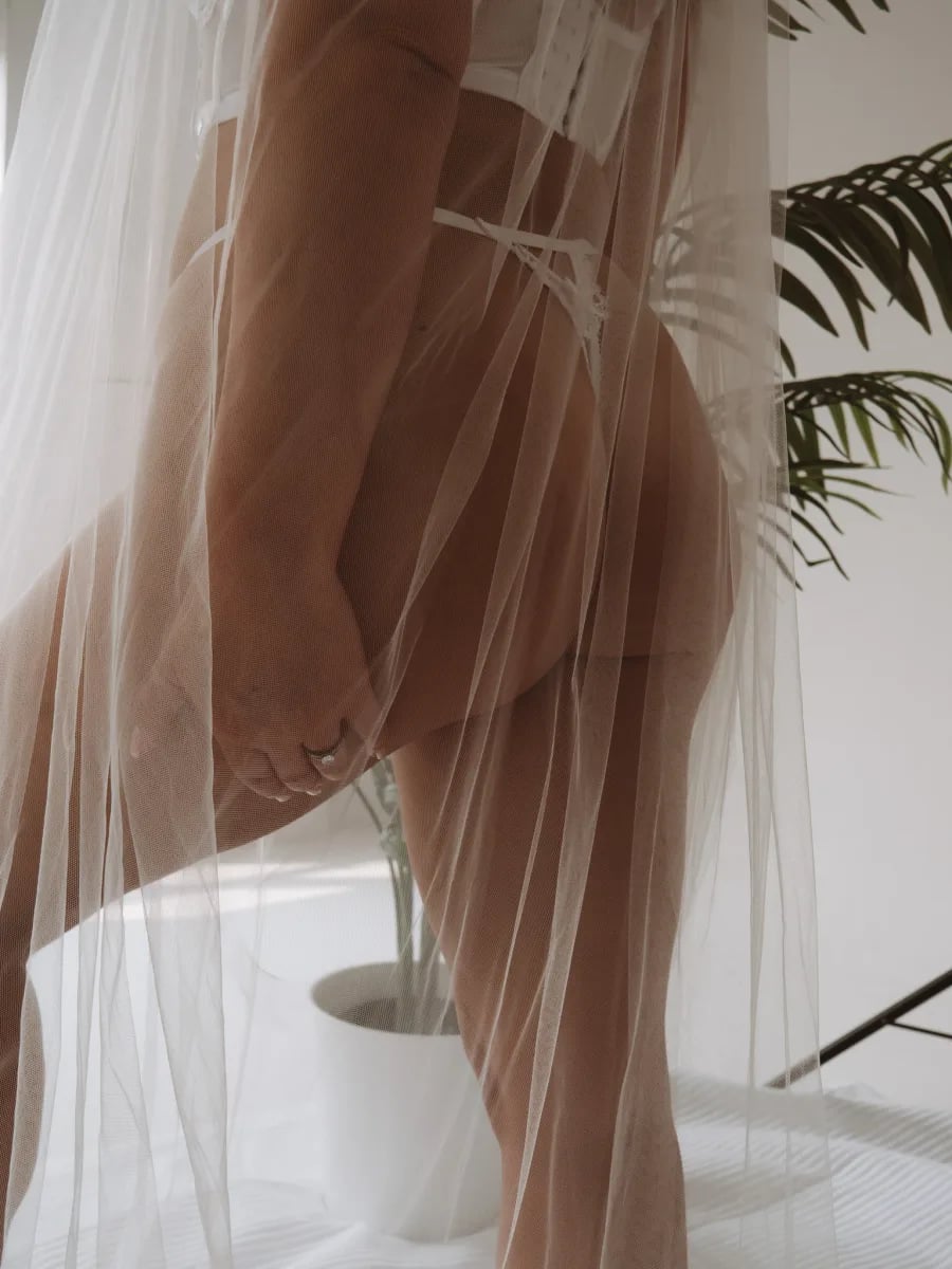 A person wearing a sheer tulle veil or fabric draped around their shoulders and torso, standing indoors with potted plants visible in the background.