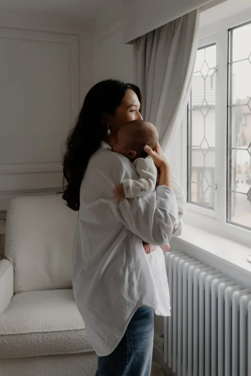 Woman in white cardigan holding a baby against her shoulder, looking out of a window with grey curtains