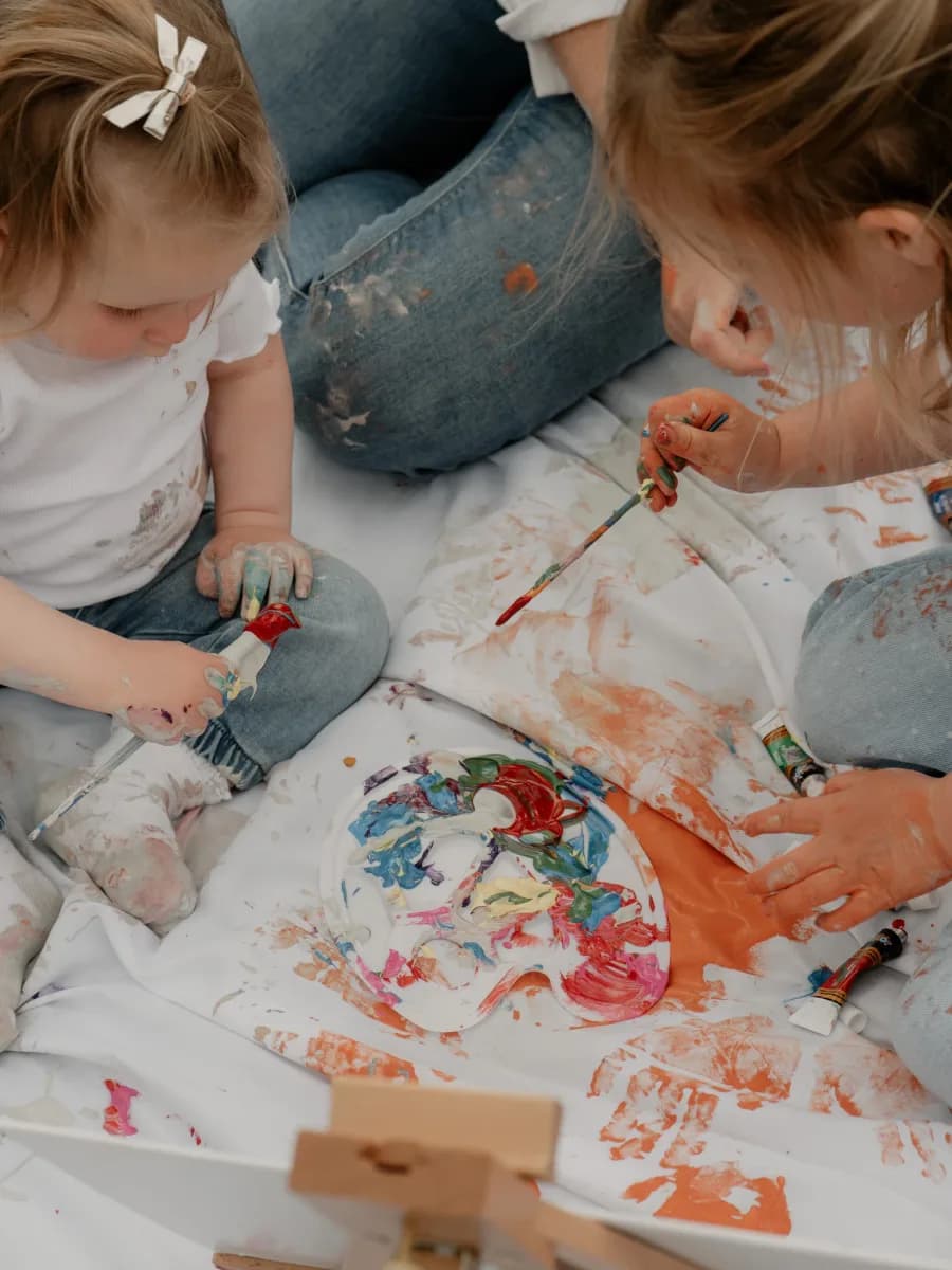 Two young children sitting on a white sheet covered in paint, creating artwork together with their hands and paintbrushes