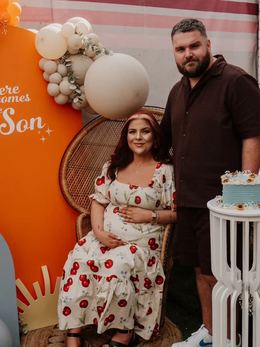A woman in a red and white floral dress and a man in a dark shirt stand together in front of an orange banner with cream and white balloons and a woven circular backdrop, with a tiered cake visible to the right.