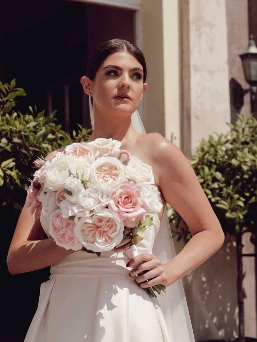 Woman in a white wedding dress holding a large bouquet of white and pink roses, standing outdoors against a pale stone wall