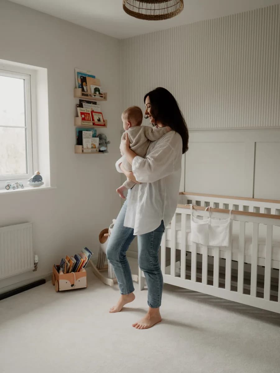 A woman holding a baby in a nursery room with a white cot and shelving on the wall