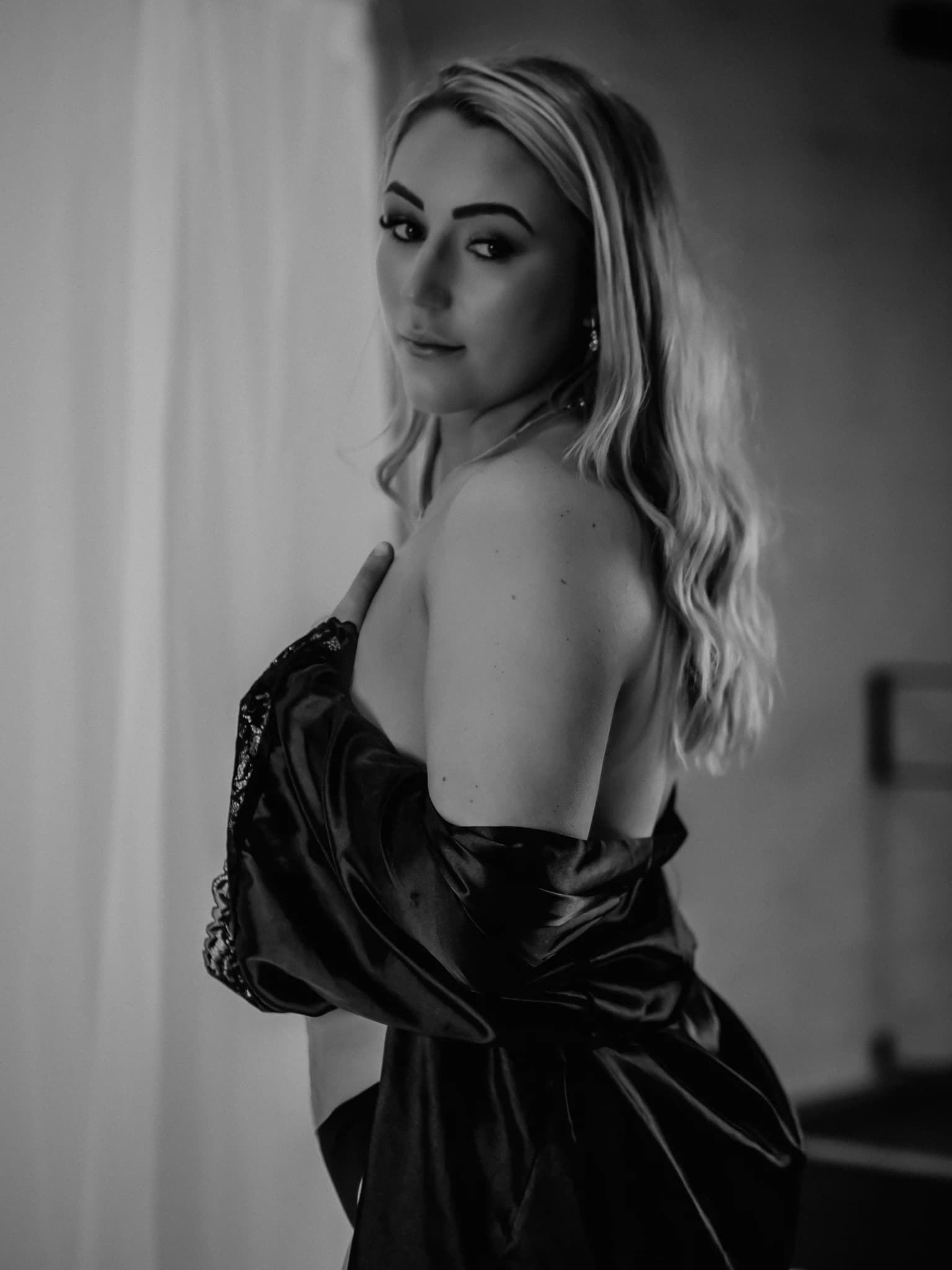 Woman with long blonde hair wearing a black silk garment, standing and looking towards the camera over her shoulder