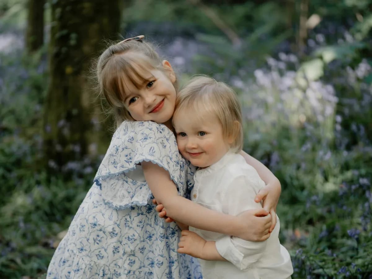 Two young children embrace outdoors amongst greenery, both smiling at the camera