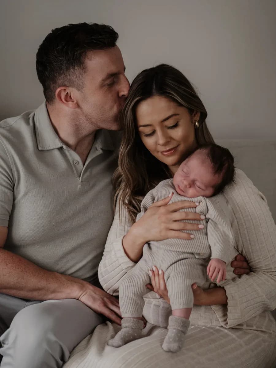 A man and woman sit together holding a newborn baby dressed in pink and white clothing