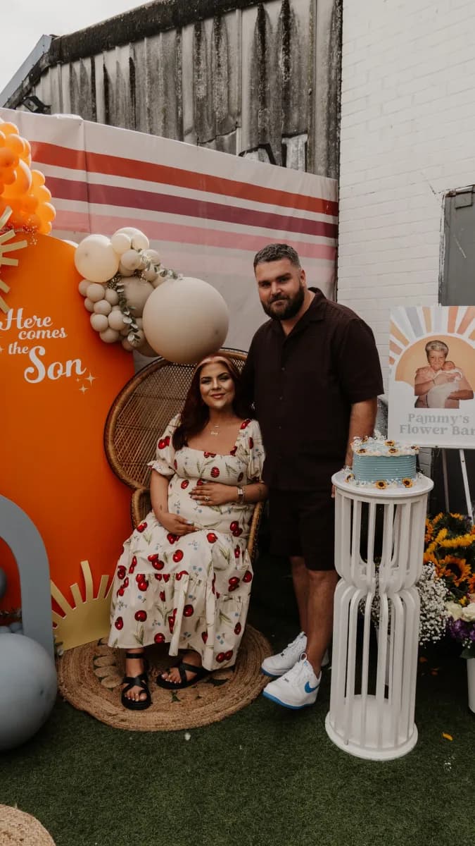 A man and woman stand together in front of a decorated backdrop with red and white stripes, surrounded by props including a large cream-coloured balloon and wooden structures.