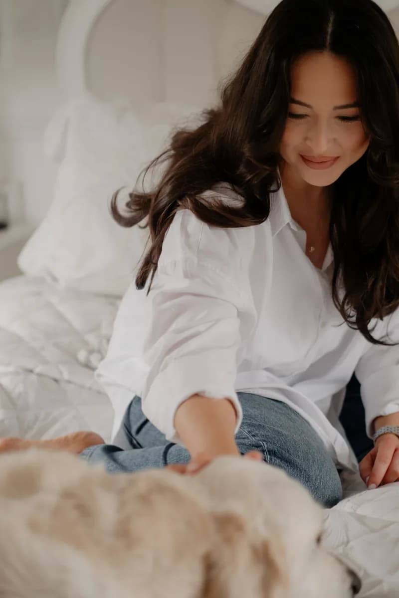 A woman in a white shirt and jeans sits on a bed, looking down at a baby in light-coloured bedding