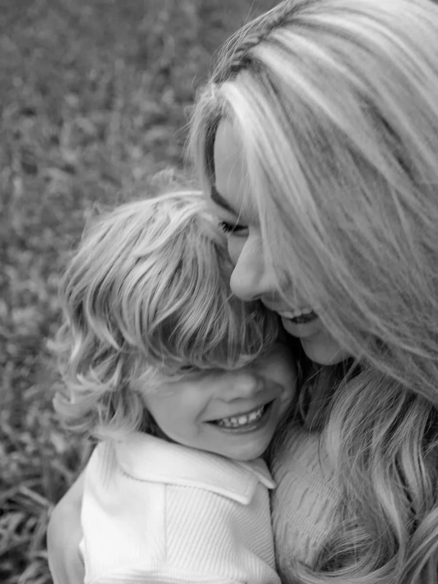 A woman with long hair hugs a young child from behind, both smiling at the camera in black and white