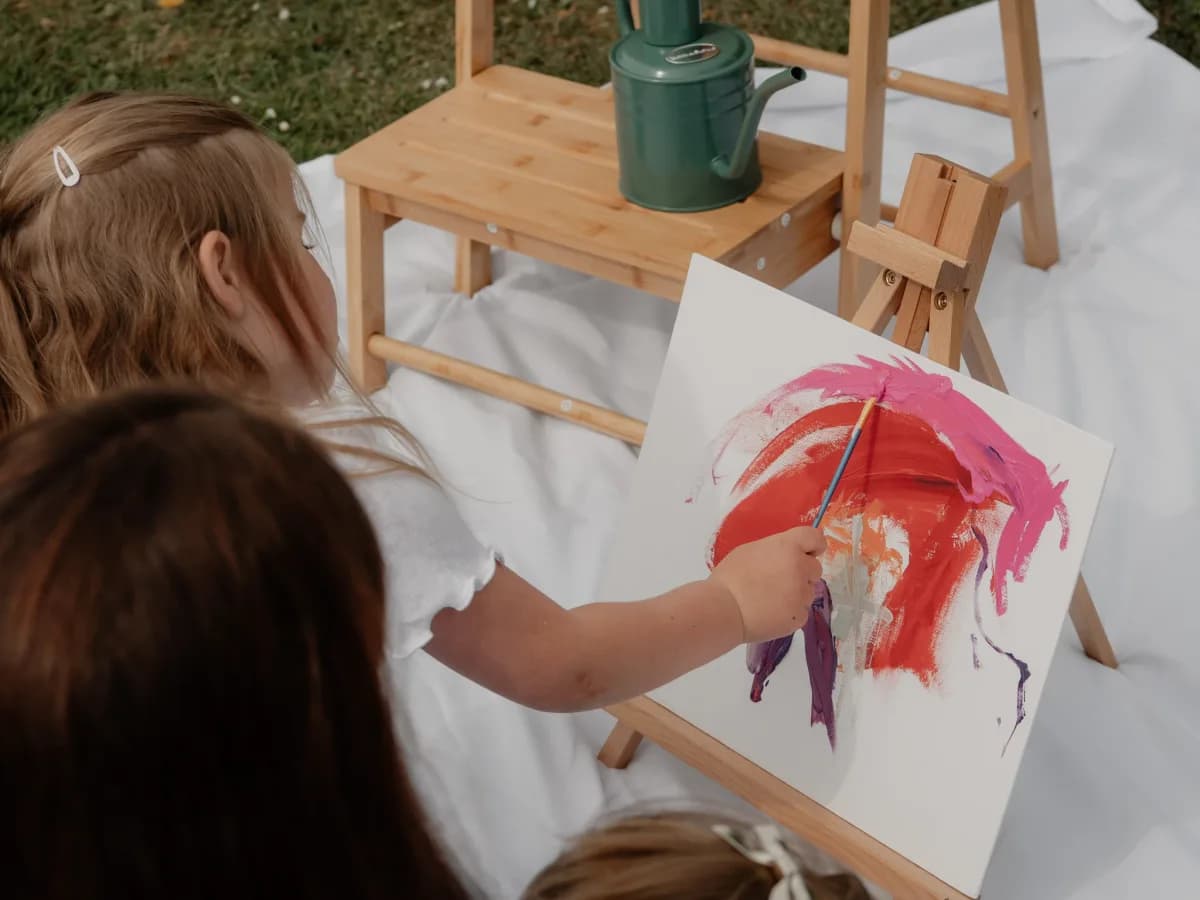 A child painting with red and pink paint on white paper at a wooden easel outdoors, viewed from behind.