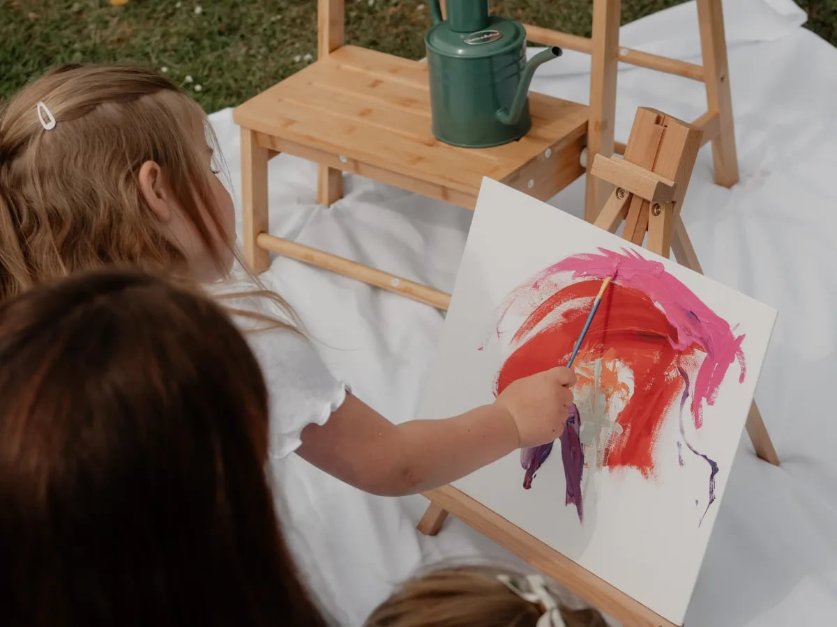A child painting with red and pink paint on white paper at a wooden easel outdoors, viewed from behind.