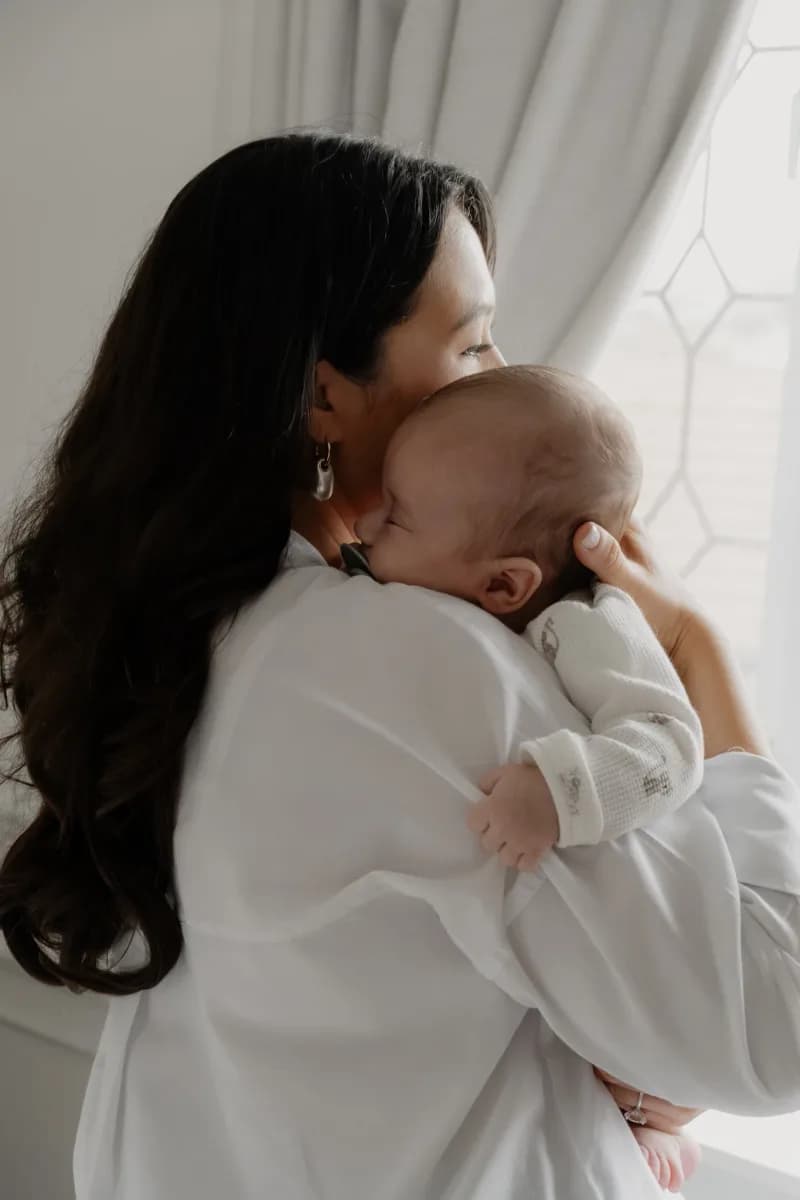 Woman with long dark hair wearing a white shirt holds a baby against her shoulder in front of a window with white curtains