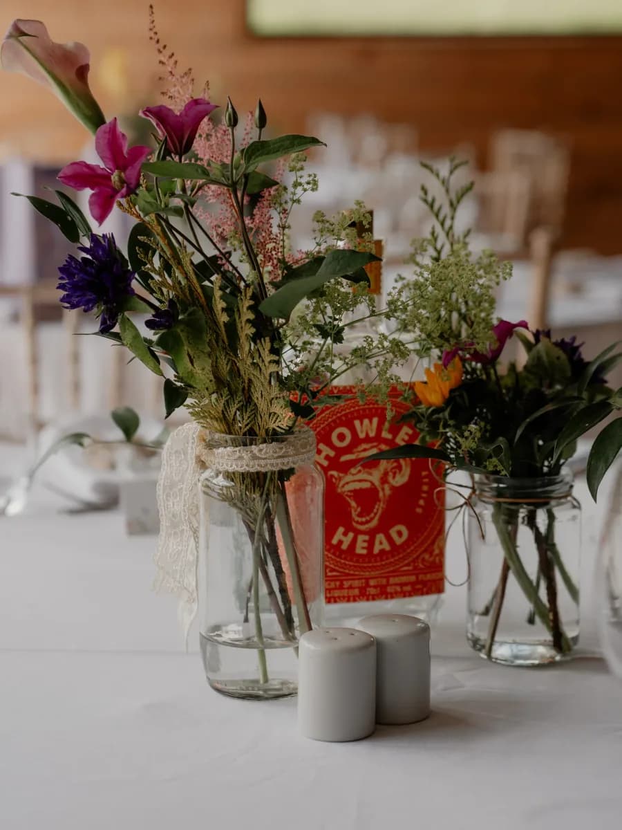 Floral centrepieces on a white table featuring clear glass vases with pink and purple flowers, a red bread bag, and grey candles in soft focus background
