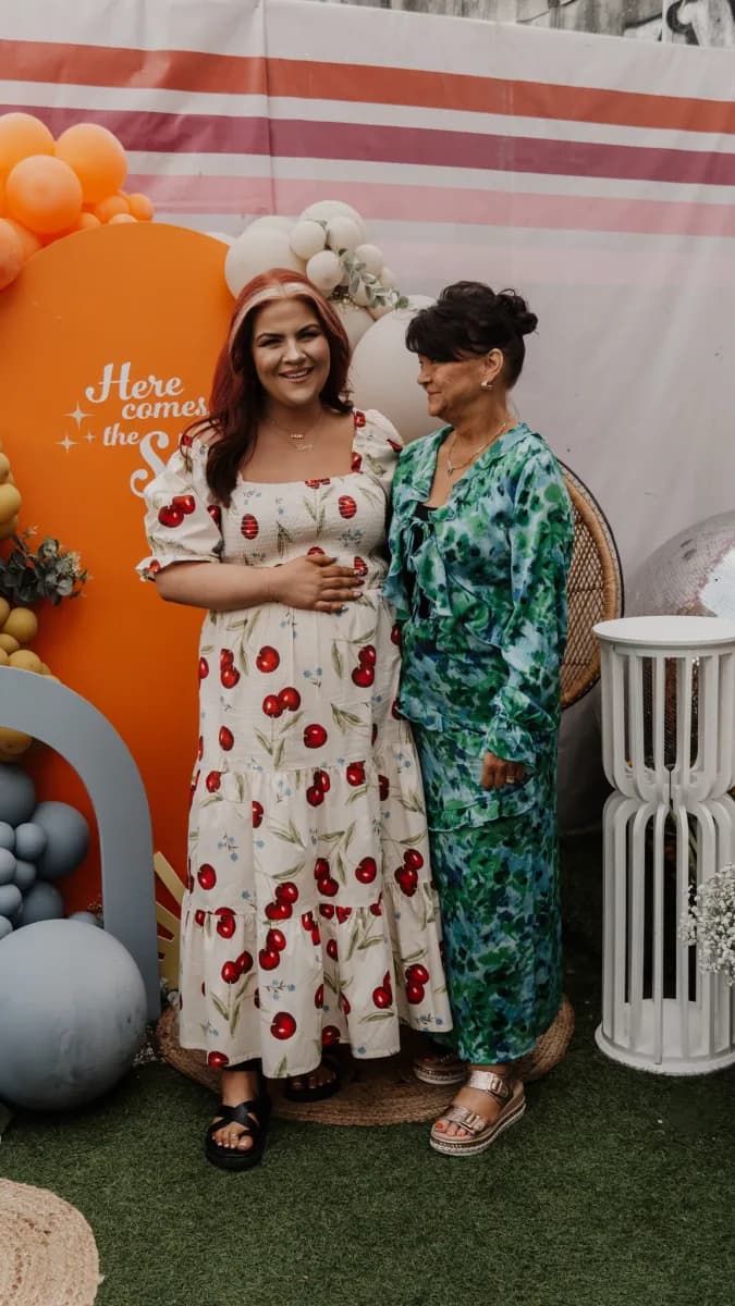 Two women standing together in front of a striped backdrop with orange balloons and white flowers, one wearing a cream dress with red pattern and the other wearing a green floral dress