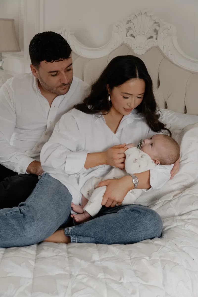 A man and woman sit together on a bed whilst the woman holds a baby in white clothing
