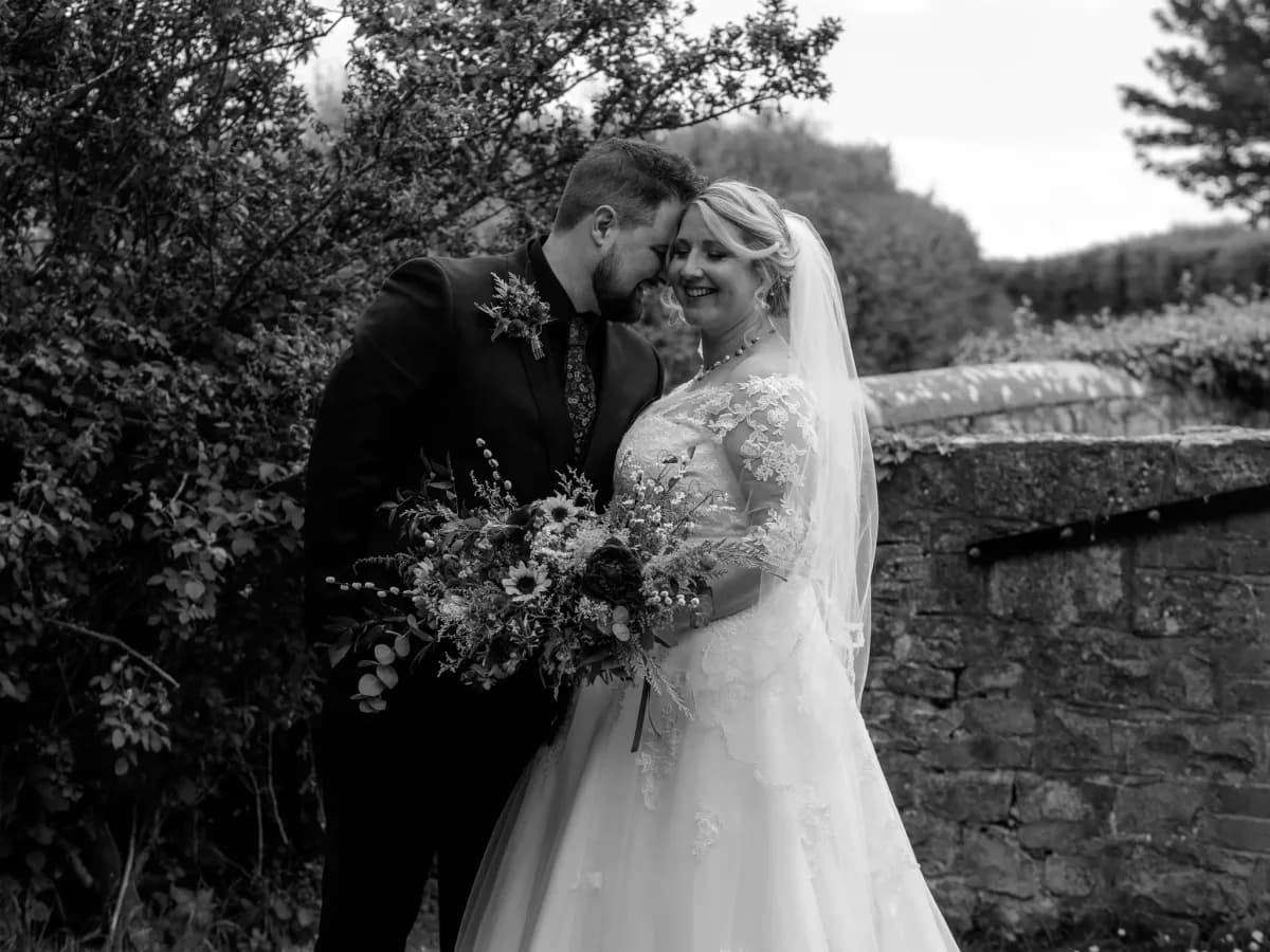 Two people in wedding attire embracing beside foliage and a stone wall in a garden setting