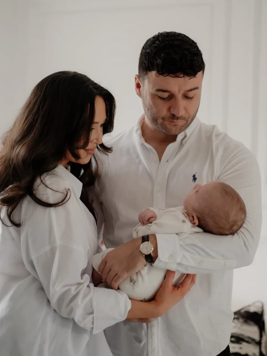 A man and woman in white shirts holding a newborn baby wrapped in white clothing