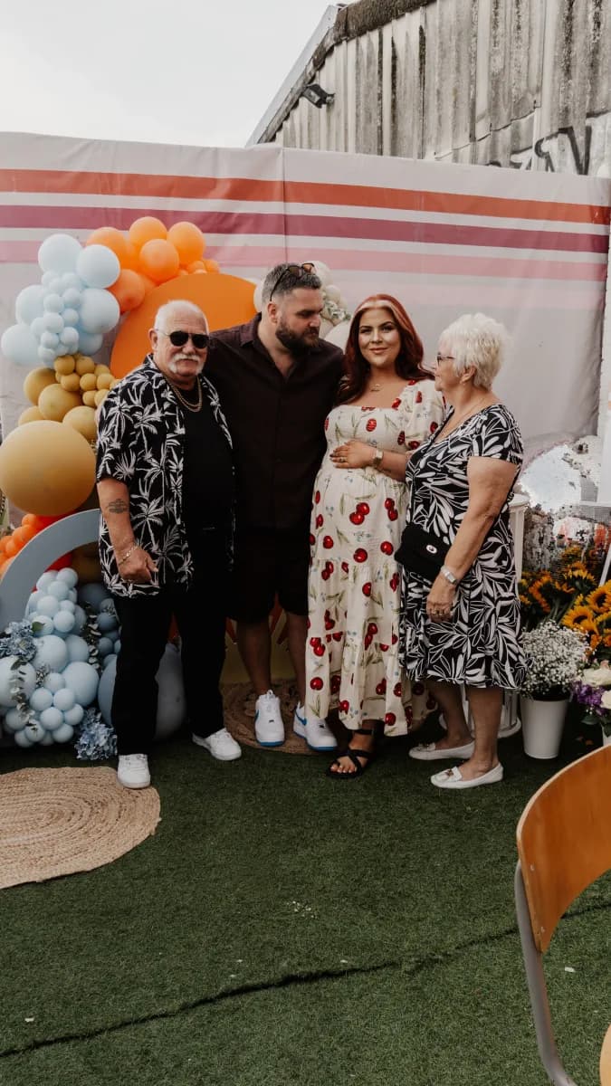 Four people stand together in front of a striped backdrop decorated with coloured balloons, on artificial grass