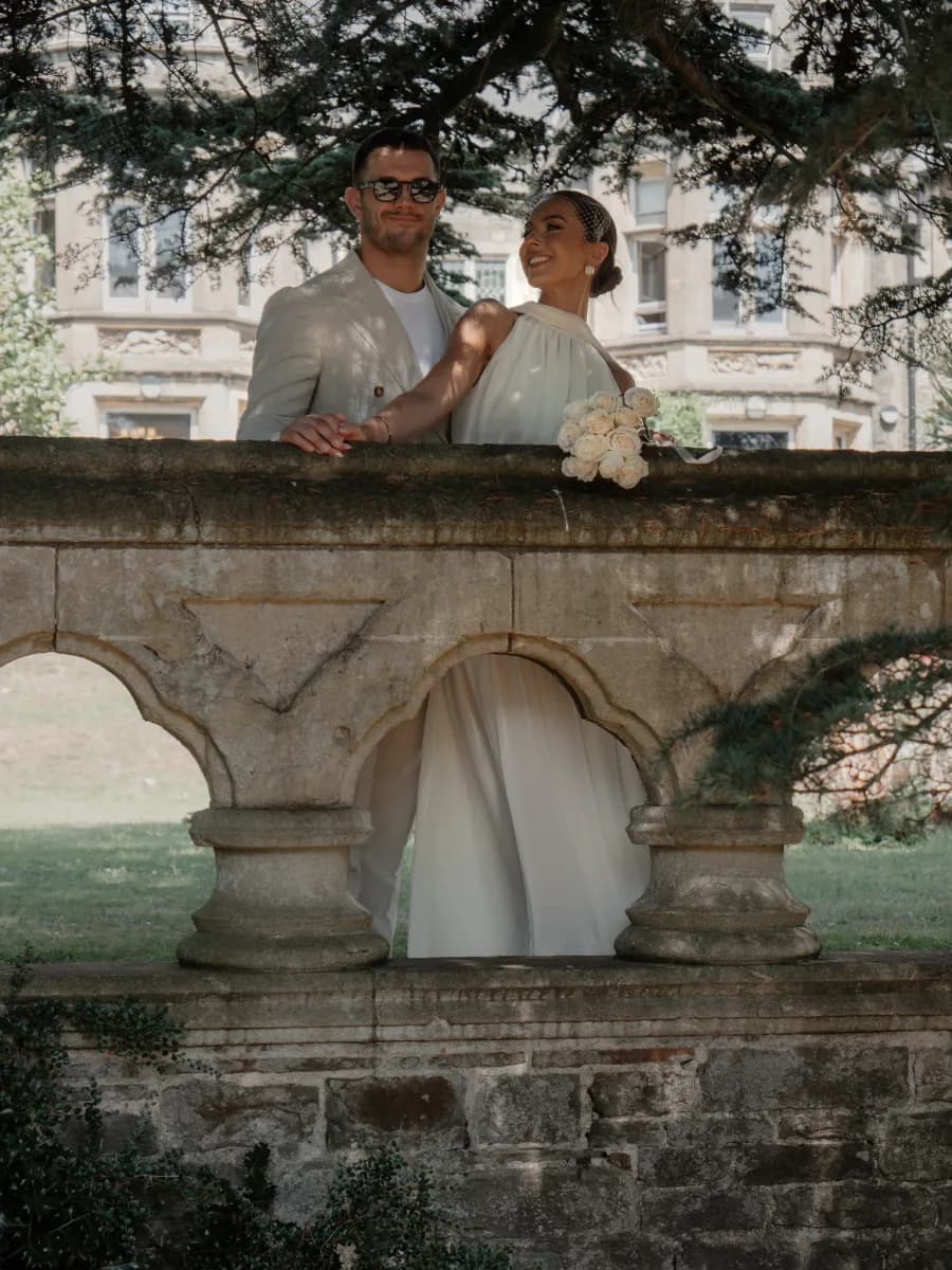 A man and woman stand together on a stone bridge with arched supports below them, surrounded by overhanging tree branches