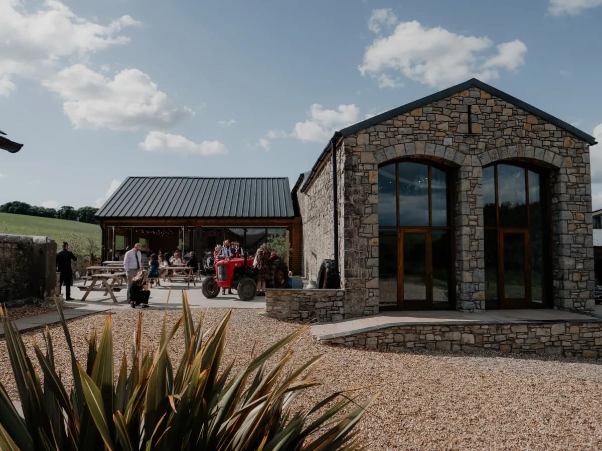 A stone building with large windows stands on a gravel courtyard, with people gathered on a patio area to the left and green fields visible in the background