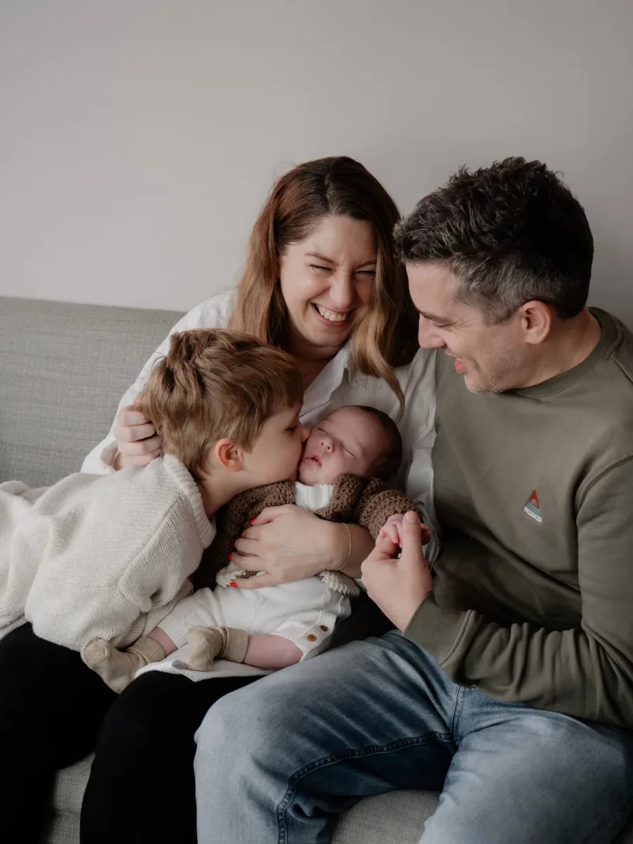 Two adults and two children sitting together on a grey sofa, all looking down at the baby being held in the centre
