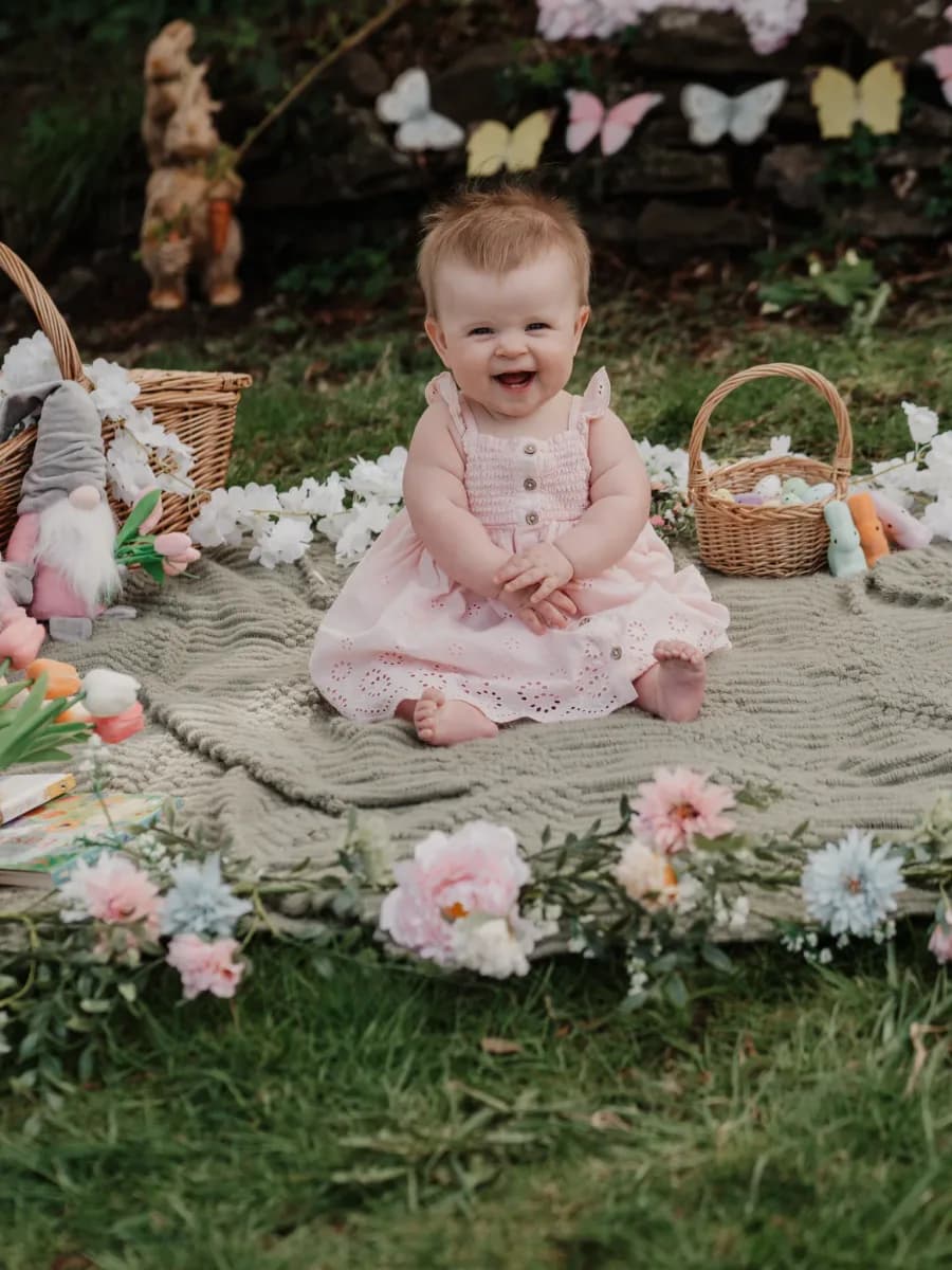 A baby in a pink dress sits on a blanket surrounded by flowers, baskets and decorative butterflies in a garden setting