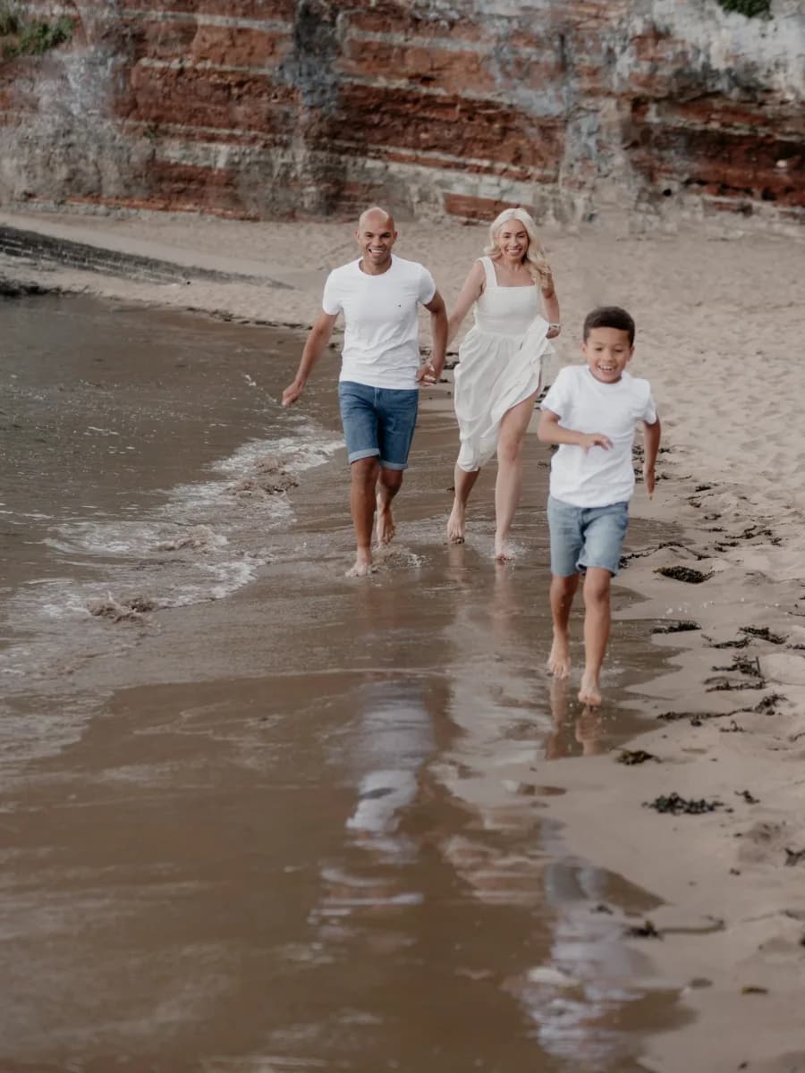 Three people running through shallow water on a beach with a brick wall behind them